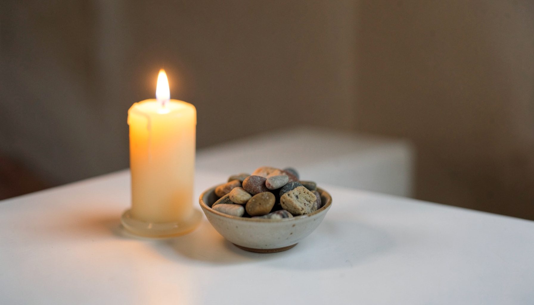 A simple still life of a candle glowing beside a small bowl of stones on a white table, warm and contemplative.